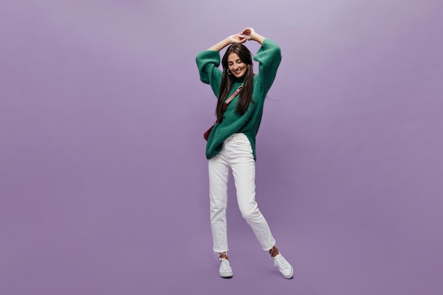 Портрет привлекательной женщины в белых брюках и зеленом свитере в полный рост Счастливая девушка танцует и улыбается на фиолетовом изолированном фоне