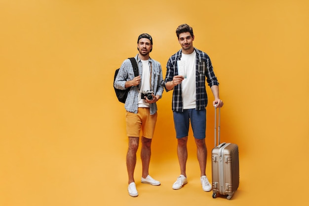 Fulllength portrait of brunet handsome men in colorful shorts checkered shirts and white tshirts hold suitcase tickets and retro camera Travelers pose on orange background