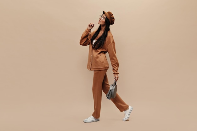 Free photo fulllength portrait of brunette longhaired woman in stylish suit moves on isolated charming girl in cap and eyeglasses holds grey handbag