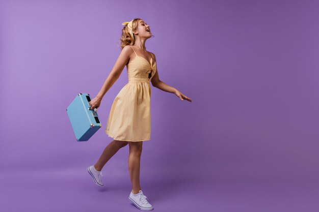 Fulllength portrait of charming white girl holding her luggage Studio shot of carefree curly lady enjoying photoshoot on violet background