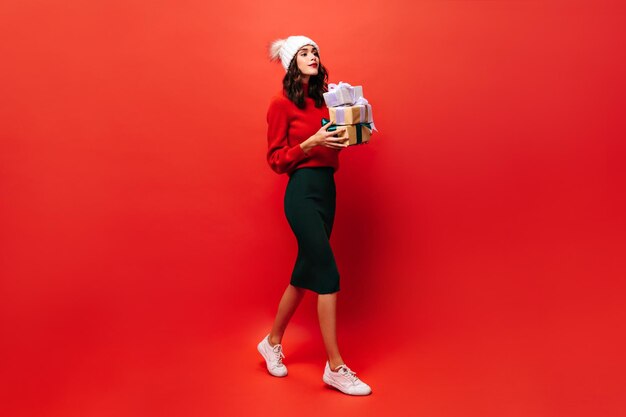 Fulllength portrait of girl holding gift box Charming woman in black skirt and bright sweater walks on red background