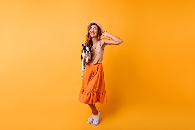 彼女の犬と一緒に時間を過ごすオレンジ色のスカートの壮大な女の子のフルレングスの肖像画黒のブルドッグでポーズをとってポジティブな笑顔の女性の屋内ショット