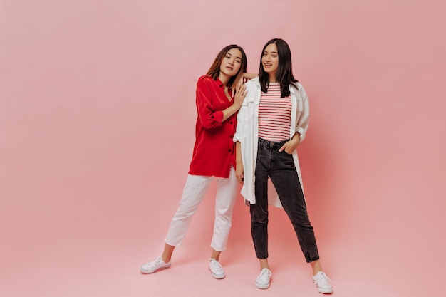 ピンクの背景に2人のアジア人女性の全身像白いズボンの短い髪の少女赤いシャツと黒のジーンズのブルネットの女性が孤立したポーズ
