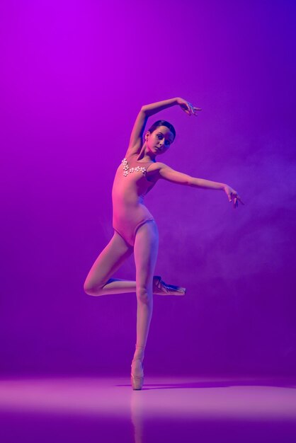 Fulllength portrait of young ballerina in pointe standing dancing isolated over gradient purple blue background in neon