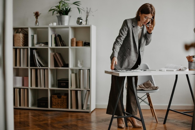 흰색 사무실에서 전화 통화를 하는 세련된 정장을 입은 젊은 비즈니스 여성의 전신 초상화