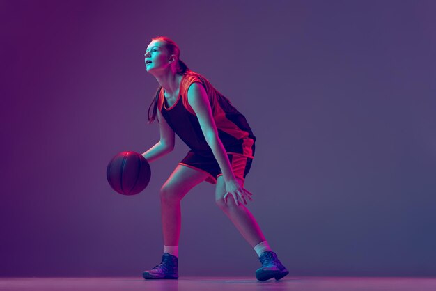 Fulllength portrait of young sportive girl basketball player training dribbling ball isolated over purple background in neon