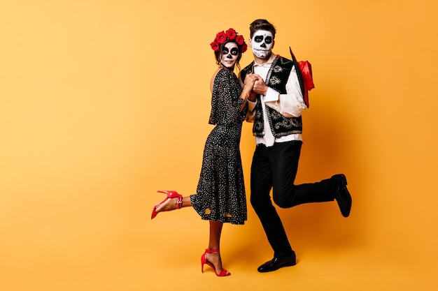 Fulllength shot of amazed couple with skeleton makeup on their faces Young people hold hands on orange background