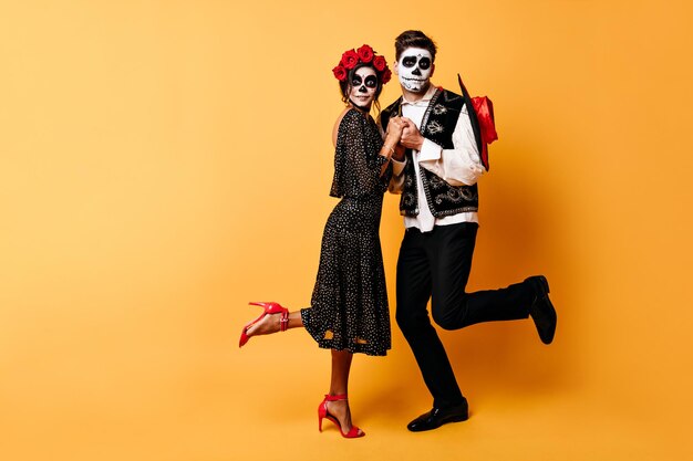 Fulllength shot of amazed couple with skeleton makeup on their faces Young people hold hands on orange background