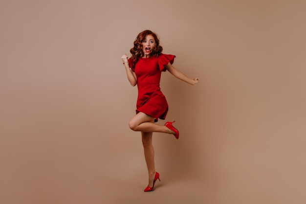 Снимок удивленной белой женщины в праздничном наряде в полный рост Потрясающая рыжая девушка в красном платье, выражающая счастье