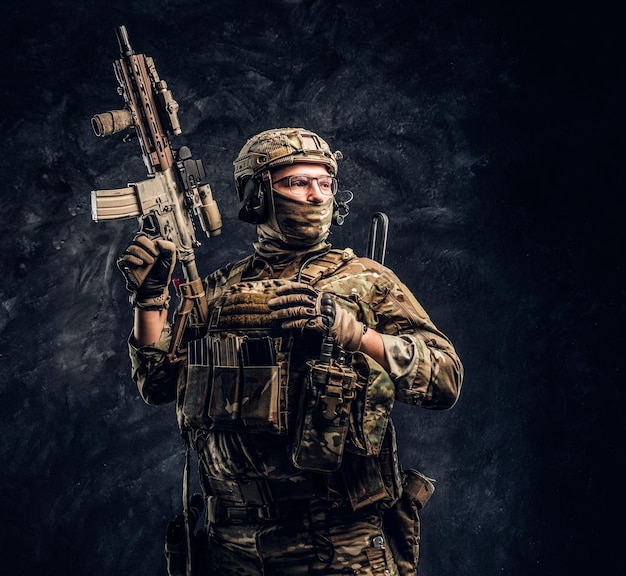 Fully equipped soldier in camouflage uniform holding an assault rifle. Studio photo against a dark textured wall