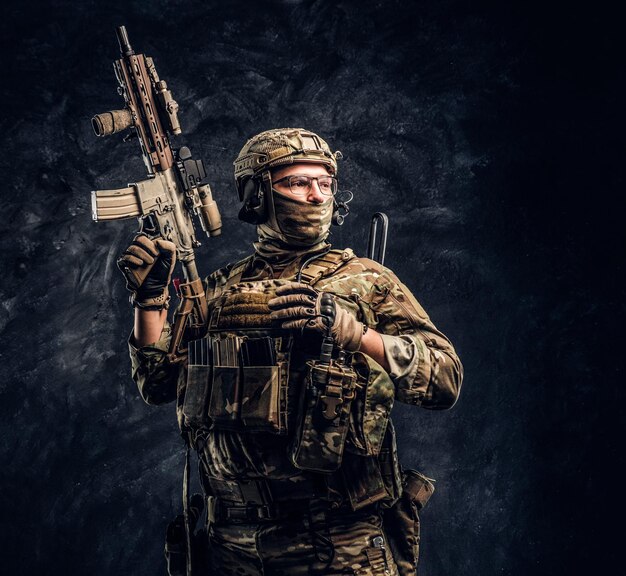 Fully equipped soldier in camouflage uniform holding an assault rifle. Studio photo against a dark textured wall