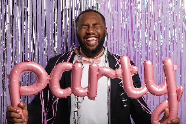 Free photo fun, happiness, holiday concept. glad dark skinned groom organises stag party, holds balloons in hands, laughs from positive emotions