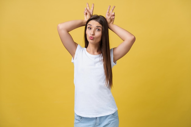 Fun and People Concept Headshot Portrait of happy Caucasian woman with freckles smiling and showing rabbit ears with fingers over head Pastel yellow studio background Copy Space