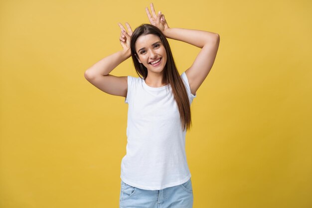 Fun and People Concept Headshot Portrait of happy Caucasian woman with freckles smiling and showing rabbit ears with fingers over head Pastel yellow studio background Copy Space