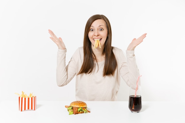 흰색 배경에 격리된 유리병에 든 감자, 감자튀김, 버거, 콜라를 입에 물고 테이블에 앉아 있는 재미있는 여자. 적절한 영양 또는 미국식 패스트 푸드. 복사 공간이 있는 영역.