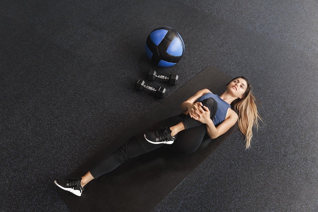 Functional training concept Attractive fit sportswoman in leggings activewear lying on rubber mat lifting one leg to stretch muscles before starting bodybuilding use med ball and dumbbells