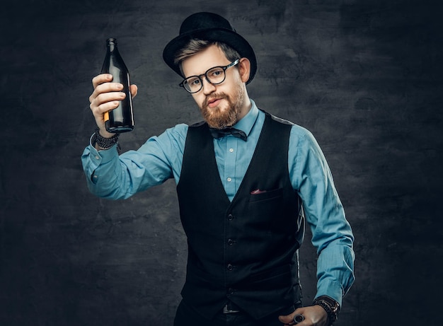 A funky bearded hipster male dressed in a blue shirt, elegant waistcoat and top hat holds a craft beer bottle.