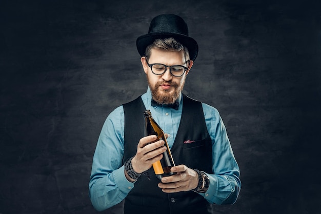 A funky bearded hipster male dressed in a blue shirt, elegant waistcoat and top hat holds a craft beer bottle.