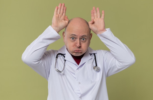 Uomo adulto divertente in uniforme da medico con lo stetoscopio che si tiene per mano sulla sua testa che gesturing le corna che guardano