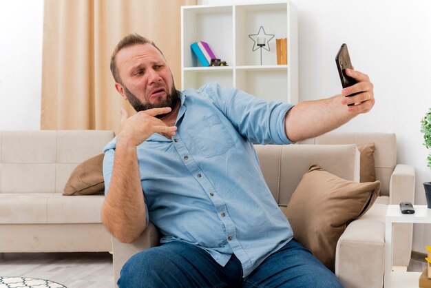Funny adult slavic man sits on armchair looking at phone taking selfie inside the living room