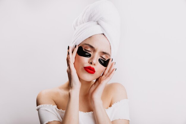 Funny adult woman in towel moisturizes skin under eyes before makeup. Lady with red lipstick poses with closed eyes on white wall.