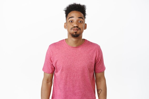 Funny african american man with afro hair and moustache, making silly pouting face and smiling, grimacing, holding breath, standing in pink t-shirt over white background.