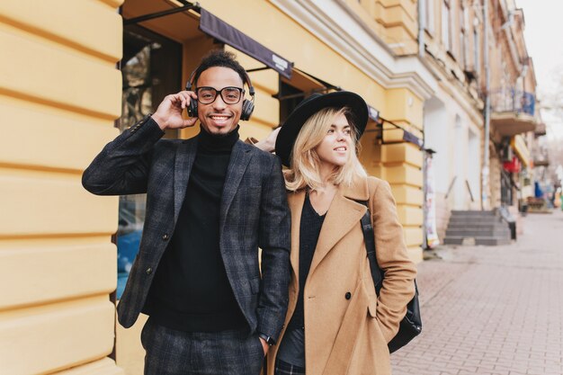 Funny african guy listening music in headphones beside lovely blonde girl. Caucasian fair-haired woman standing near smiling mulatto man in black attire.