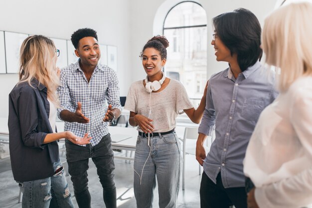 Funny african woman in vintage jeans posing between black and asian friends in international university. Freelance specialists meeting with foreign colleagues.