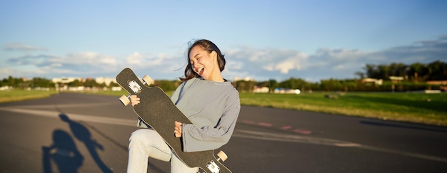 Funny asian girl enjoying skating holding skateboard like guitar and shadow playing having fun