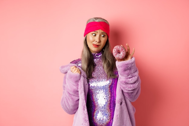 Funny asian senior woman in glittering disco dress and faux fur coat looking tempted at delicious donut, wanting to eat sweet, standing over pink background