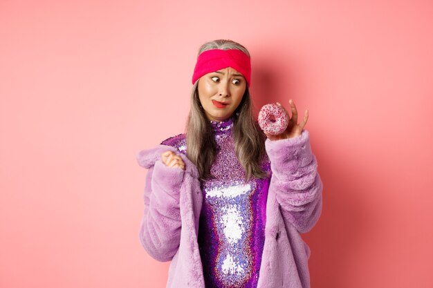 Funny asian senior woman in glittering disco dress and faux fur coat looking tempted at delicious donut, wanting to eat sweet, standing over pink background