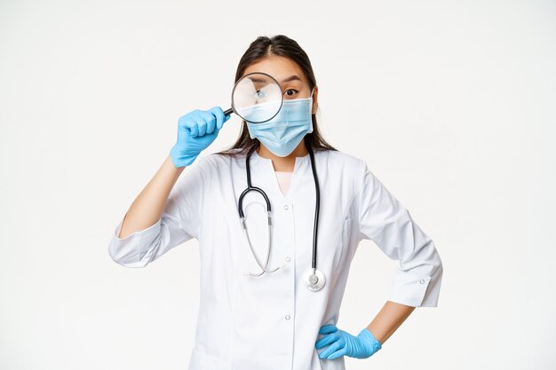 웃긴 아시아 여성 의사는 돋보기를 통해 환자를 바라보고 의료용 얼굴 마스크와 고무 장갑을 끼고 흰색 배경