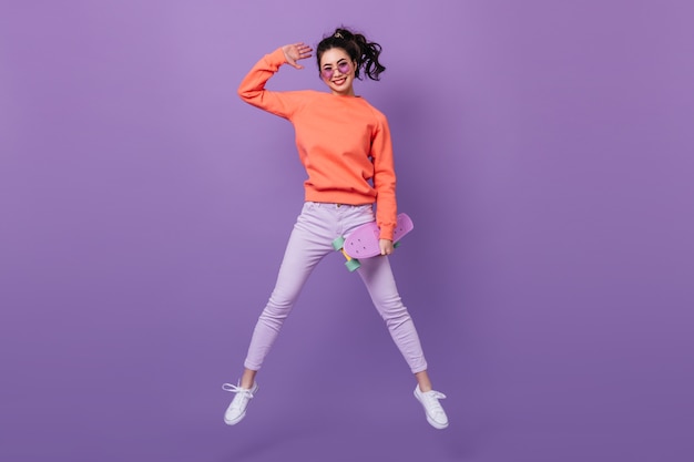Funny asian woman in pants jumping on purple background. full length view of korean young woman with longboard