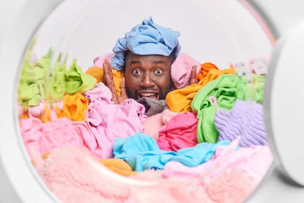 Funny bearded Afro American man covered with multicolored clothes collected for washing poses from inside of washer keeps hand raised smiles broadly