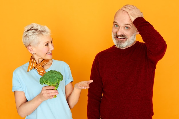 Funny bearded male pensioner holding hand on his chest having puzzled confused look does not want to eat broccoli which wife offering him.