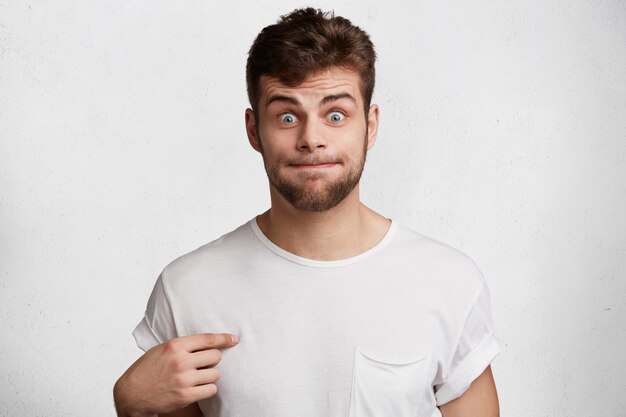 Funny bearded male presses lips, points at blank casual t shirt