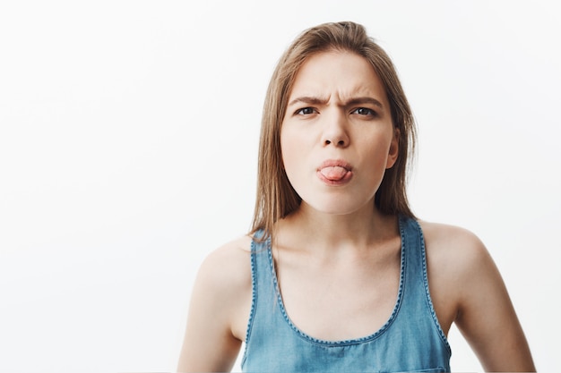 Funny beautiful dark-haired student girl in casual blue shirt showing tongue,  with silly face expression, trying make little kid in park laughing.