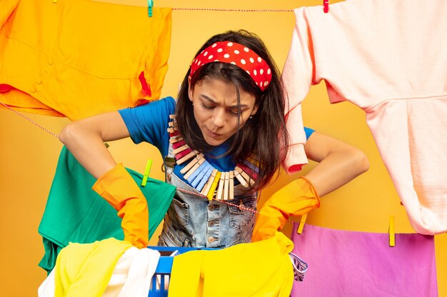 Funny and beautiful housewife doing housework isolated on yellow background. Young caucasian woman surrounded by washed clothes. Domestic life, bright artwork, housekeeping concept. Looks busy.