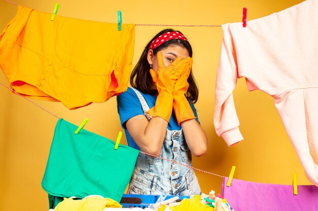 Funny and beautiful housewife doing housework isolated on yellow background. Young caucasian woman surrounded by washed clothes. Domestic life, bright artwork, housekeeping concept. Scared.