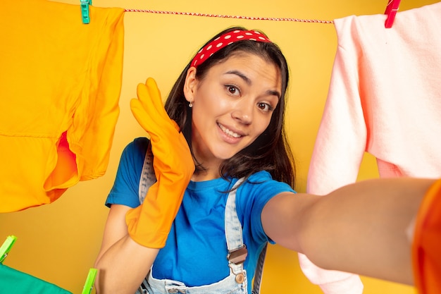 Funny and beautiful housewife doing housework isolated on yellow background. Young caucasian woman surrounded by washed clothes. Domestic life, bright artwork, housekeeping concept. Selfie view.