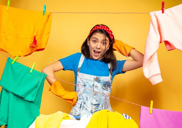 Funny and beautiful housewife doing housework isolated on yellow space. Young caucasian woman surrounded by washed clothes