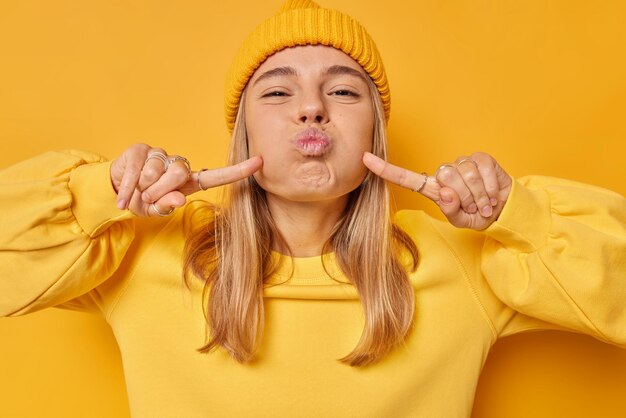 Funny beautiful young woman pouts lips blows cheeks makes grimace foolishes around wears hat casual long sleeved jumper isolated over vivid yellow background. Human face expressions concept.