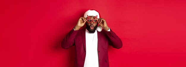 Funny black man celebrating new year wearing party glasses and santa hat having fun over red backgro