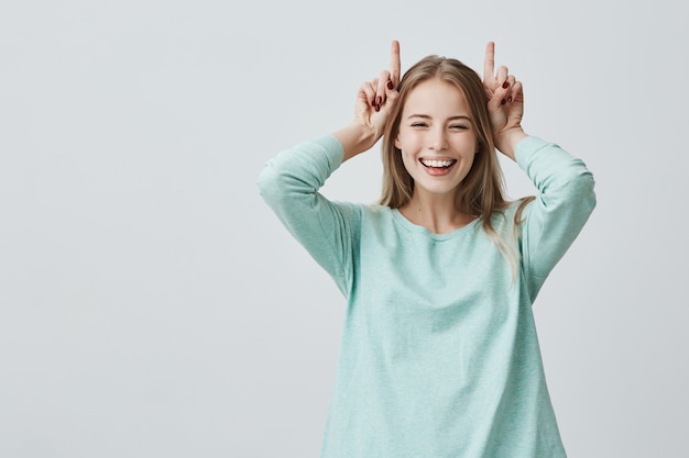 Funny blonde woman smiling broadly holding fingers above head. Horns gesture