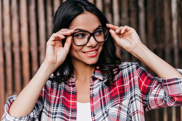 Funny brunette girl looking away and smiling. Outdoor portrait of cute happy woman in glasses wears checkered shirt.