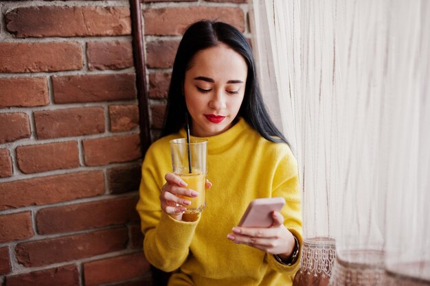 Free photo funny brunette girl in yellow sweater drinking juice and looking at mobile phone at restaurant