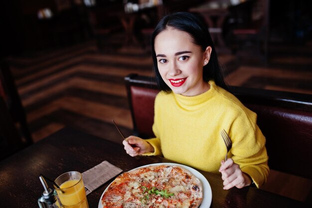 Funny brunette girl in yellow sweater eating pizza at restaurant