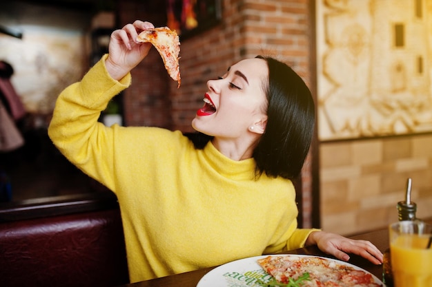 レストランでピザを食べる黄色いセーターの面白いブルネットの女の子