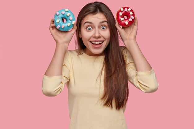 Funny brunette lady woman holds colourful doughnuts, looks happily directly 