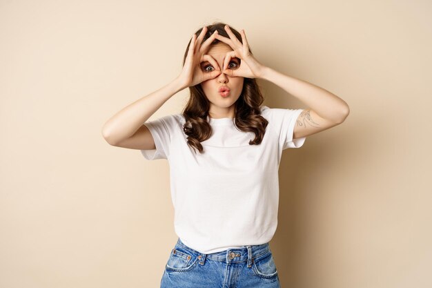 Funny brunette woman having fun, showing finger glasses gesture and fool around, posing over beige background